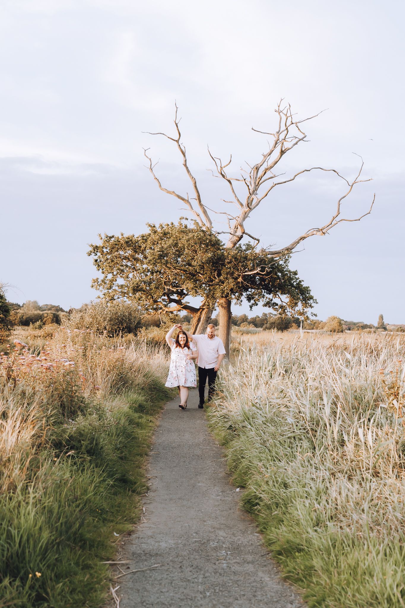 Couple walking on path through tall grass field beneath bare branched tree.