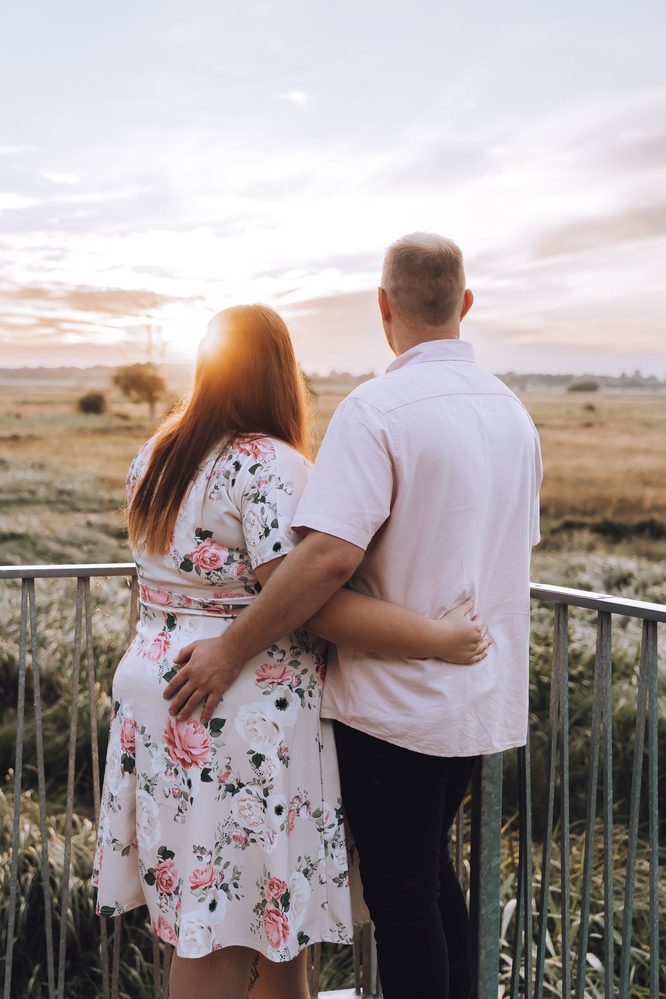 Couple embraces while watching sunset from a balcony overlooking a serene countryside landscape.