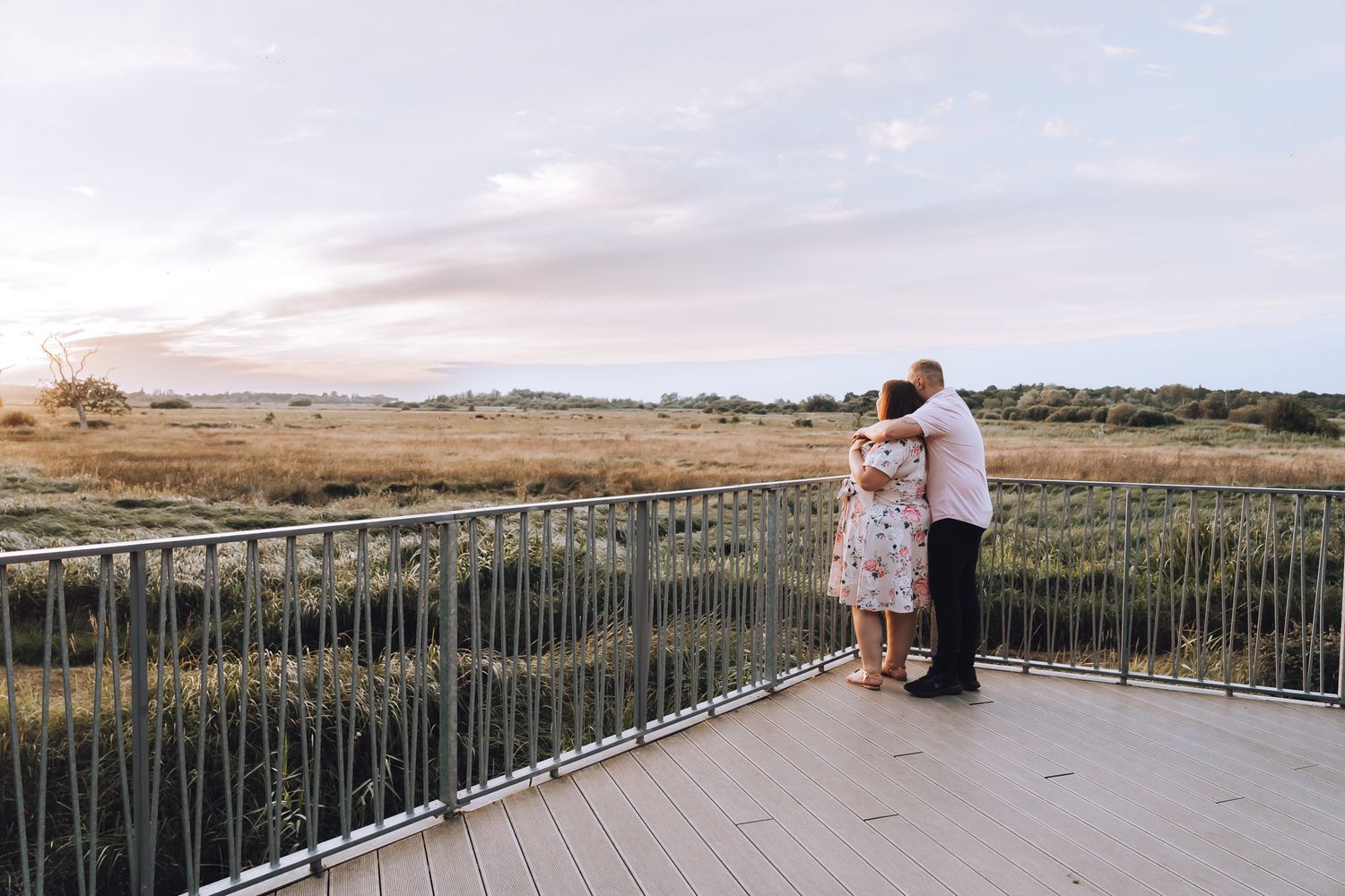 Romantic couple stands together on observation deck overlooking scenic rural landscape at sunset.