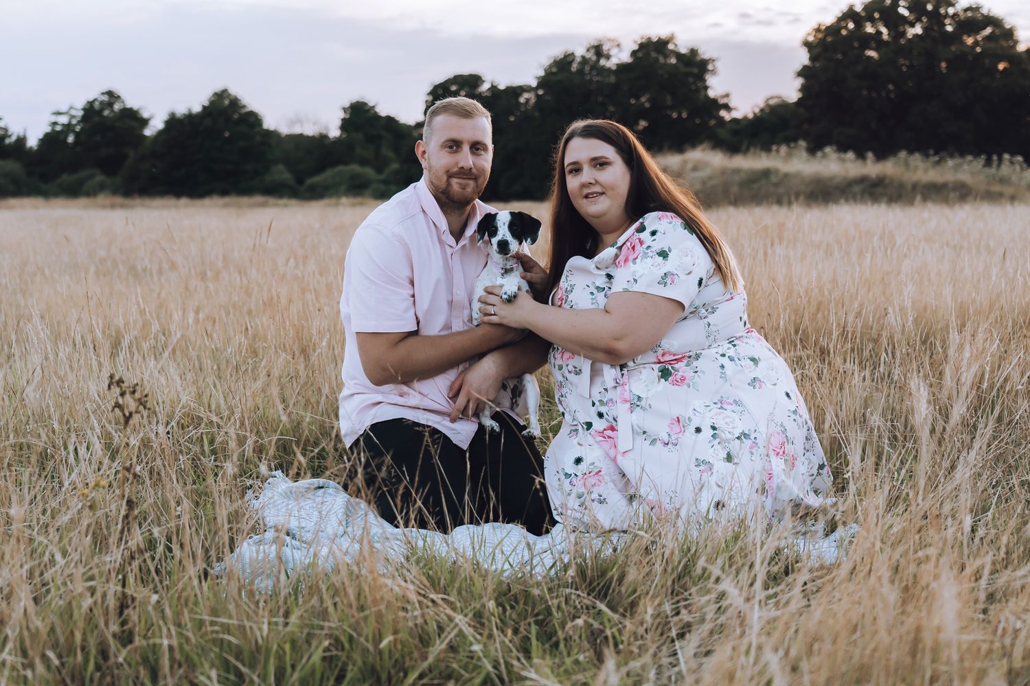 Happy couple poses with their small dog in a scenic field of tall grass at dusk.