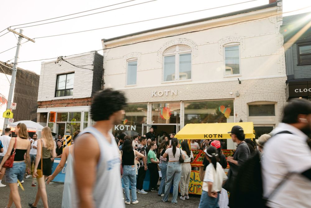 Crowd of people gathered outside urban storefronts during a daytime event.
