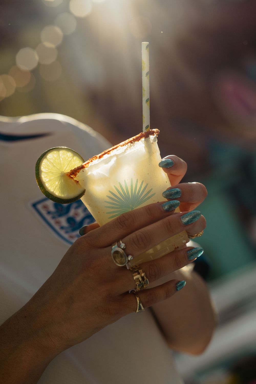 Hand with blue nails and rings holding a margarita cocktail with lime and straw in golden sunlight.