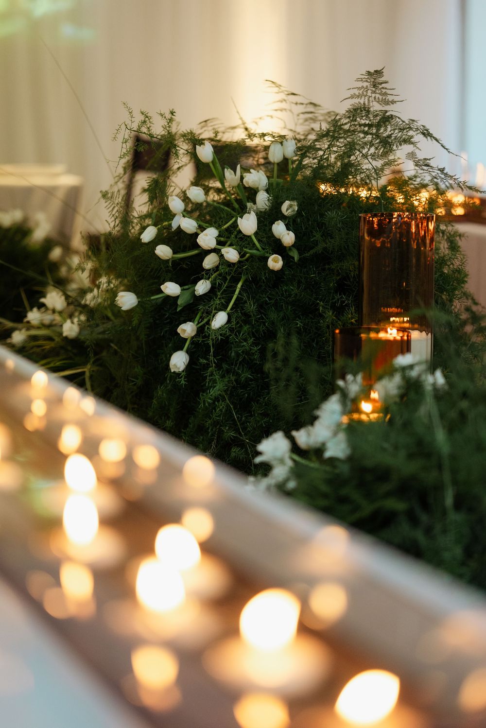 Elegant wedding table with white tulips, lush greenery, amber votive candles, and warm glowing tealights creating romantic ambiance.