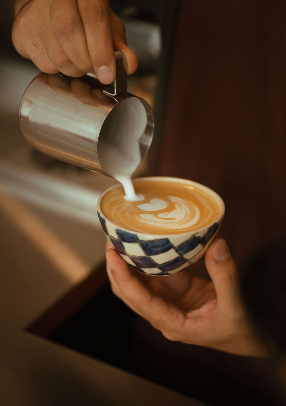A barista pours steamed milk into a latte art design in a decorative blue and white ceramic cup.