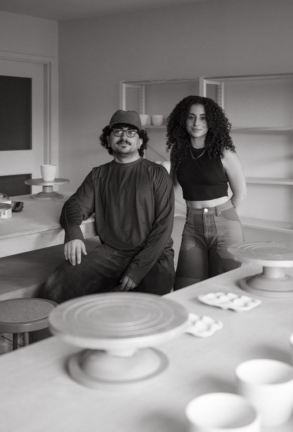 Two artists pose in a ceramic studio surrounded by pottery wheels and handcrafted pieces in black and white.