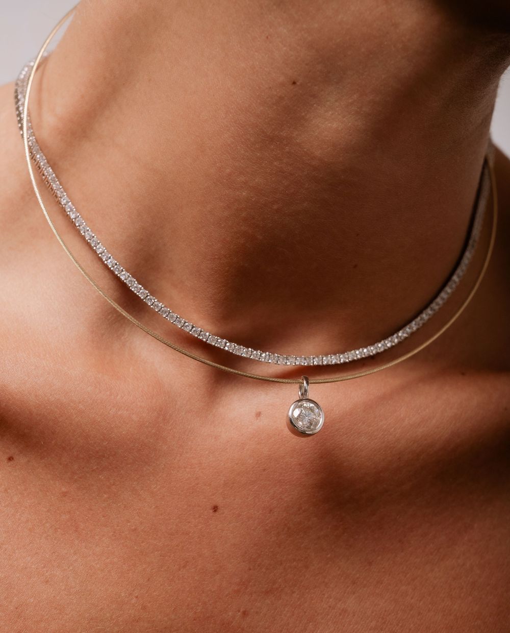 Silver tennis necklace layered with a delicate chain pendant necklace featuring a round crystal charm on a model.