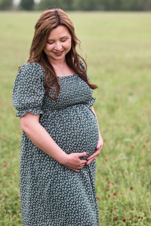 A pregnant woman in a green floral dress stands in a wildflower field cradling her baby bump.