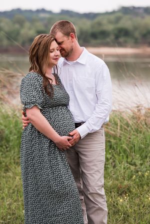 Tender maternity photography session with couple in white shirt and floral dress against natural outdoor backdrop.
