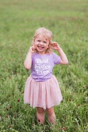 A small child wearing a purple 'Best Sister Ever' shirt plays joyfully in a grassy field.