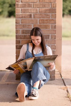 Student sits against brick wall reading books while wearing casual denim and checkered shoes.