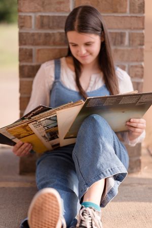 Reader in denim overalls relaxes with books against brick wall wearing sneakers.