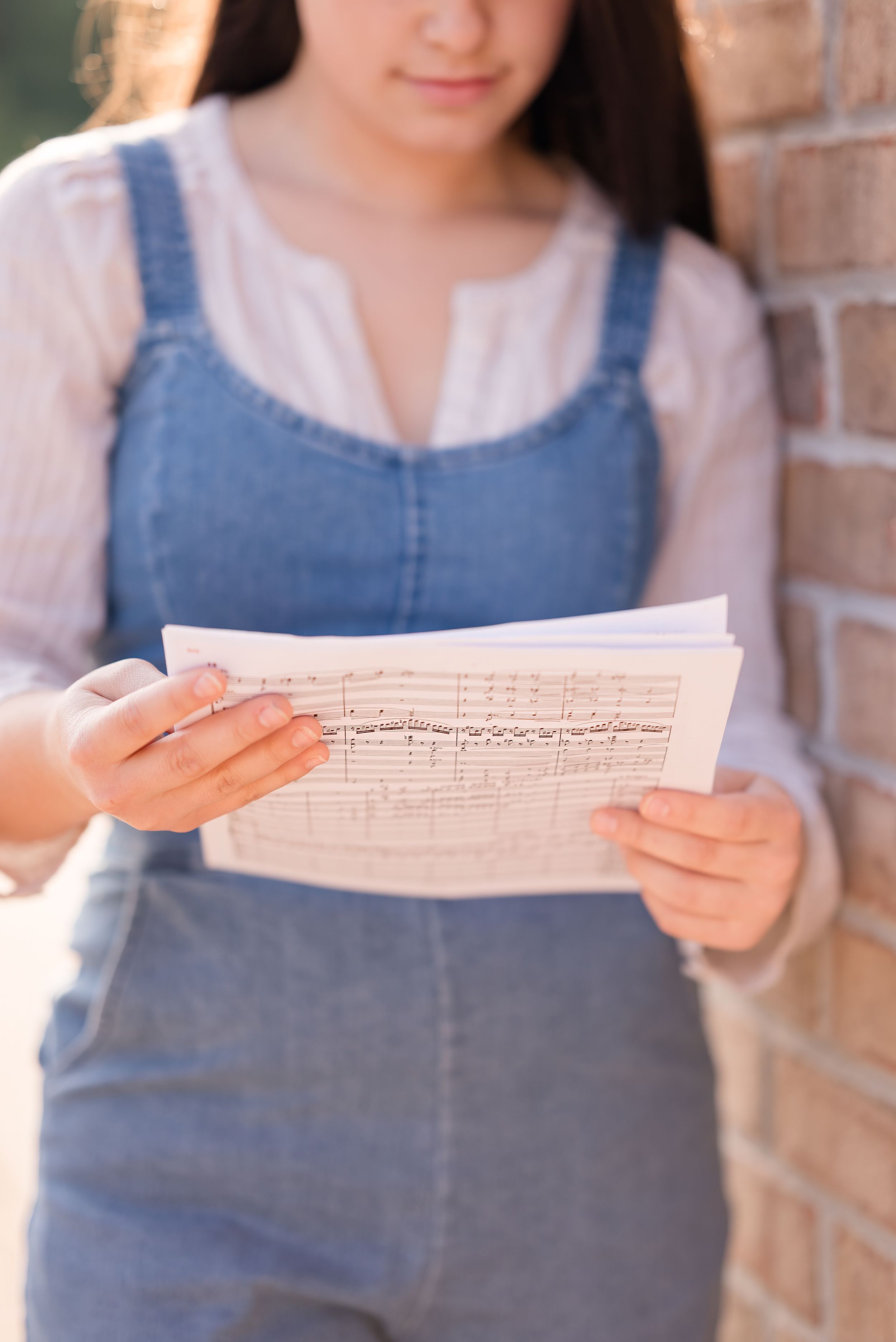 Student in denim overalls holds sheet music while standing against brick wall.