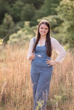 A person in denim overalls and pink top poses in a golden field during sunset.