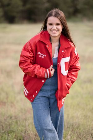 Someone wearing denim overalls and a bright red jacket poses playfully in a grassy field.
