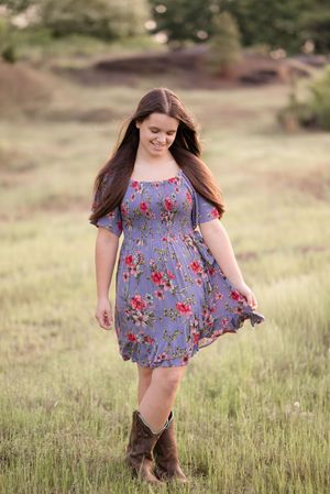 A young person in a blue floral dress and brown boots runs through a sunlit meadow of tall grass.