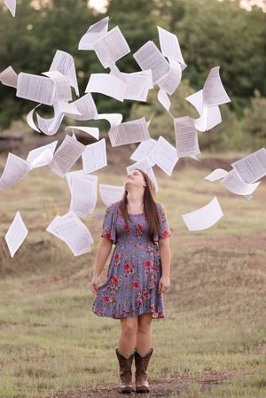 Papers flutter through the air in a grassy field as someone in a blue floral dress stands beneath them.