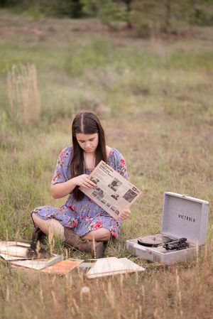 A person in a floral dress sits in a grassy field with a vintage typewriter and papers laid out on the ground.