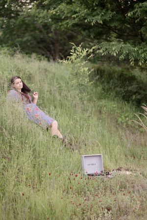 Person relaxes in a wildflower meadow with green trees and scattered red flowers in the background.