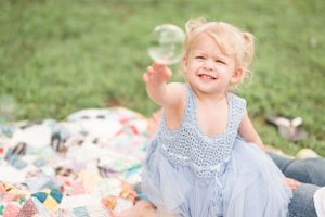 A blonde toddler in a blue dress smiles joyfully while sitting on a colorful blanket outdoors.