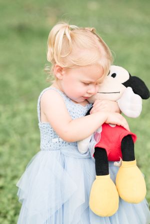 A toddler in blue dress hugging a Mickey Mouse plush toy outdoors.