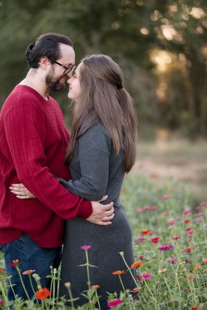 A couple embraces romantically in a blooming wildflower field at sunset wearing red and grey sweaters.
