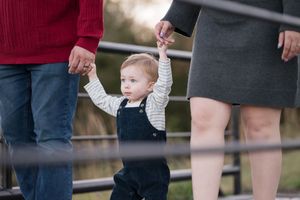 Small child in overalls holding hands with adults while learning to walk.