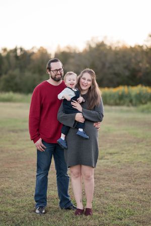 Family of three in coordinating red and gray outfits pose together in an outdoor field at sunset.