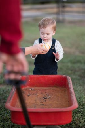 A toddler in navy overalls reaches toward a red sandbox while being helped by an adult's hand.