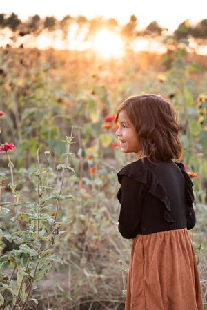 A figure in a brown dress and black cardigan stands among wildflowers during golden hour at sunset.
