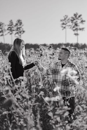 Couple stands together in a dreamy field of white wildflowers with tall pine trees in the background.