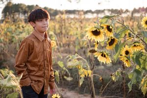 Person in brown jacket stands among blooming sunflowers in a golden autumn field at sunset.
