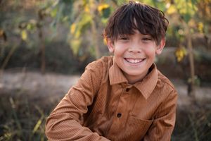 A young person wearing a brown corduroy shirt smiles brightly outdoors with autumn foliage in the background.