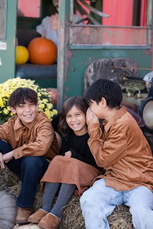 Three friends share a joyful moment while sitting on hay bales with autumn pumpkin decorations in the background.