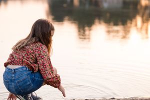 A woman in a floral dress and jeans sits by a tranquil lake during sunset, creating a peaceful reflection on the water.