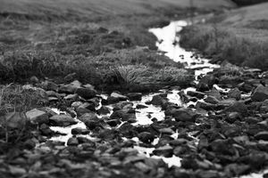 Black and white landscape photograph shows a winding stream cutting through rocky terrain in dramatic lighting.