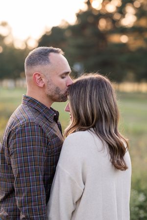 A couple shares a romantic moment in a field at sunset, wearing plaid and cream colored clothing.