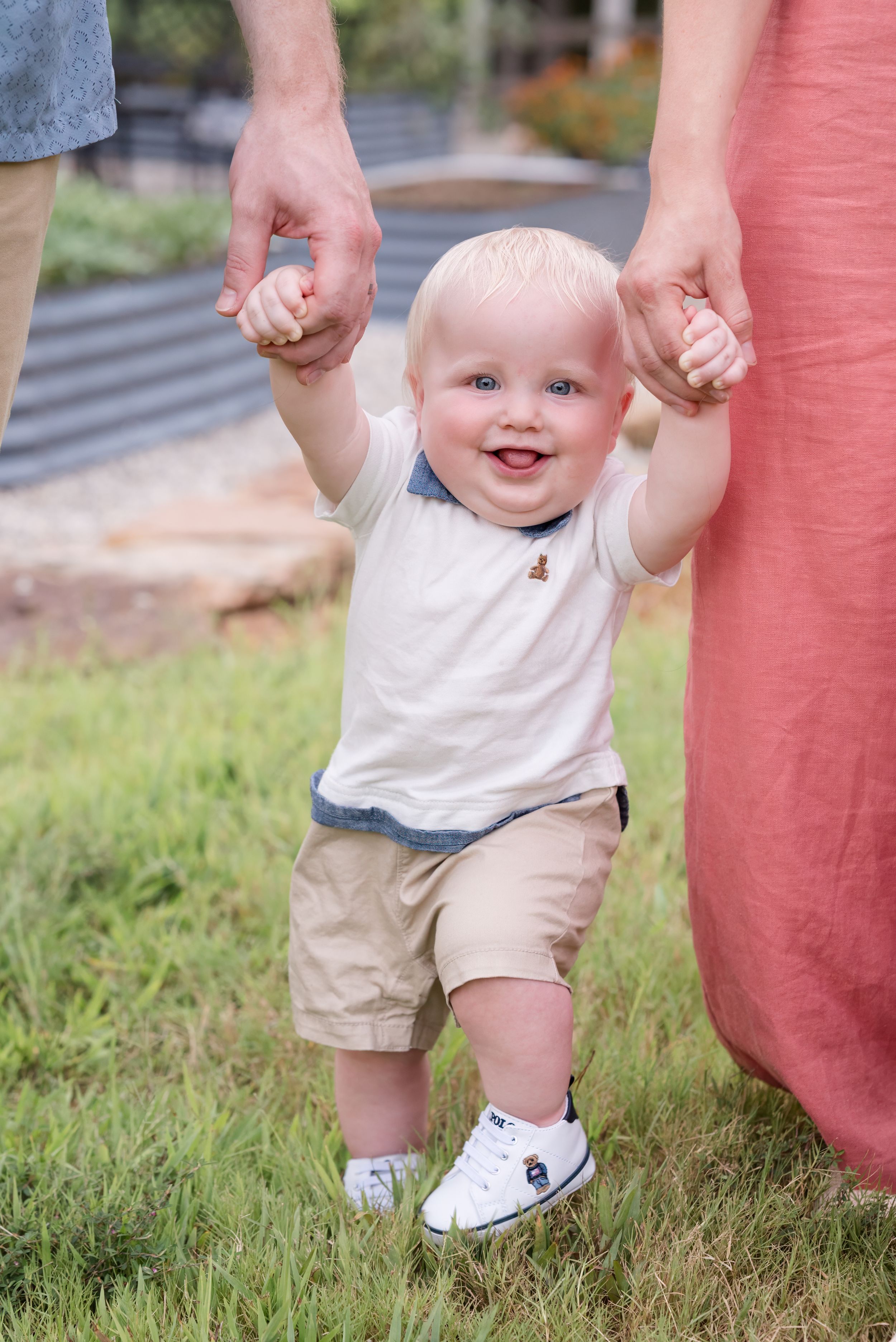 A toddler in a white shirt and khaki shorts walks while holding hands with adults on a grassy field.