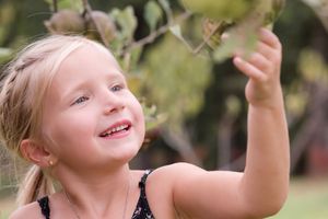 A child in a black tank top reaches up and smiles while playing outdoors on a sunny day.
