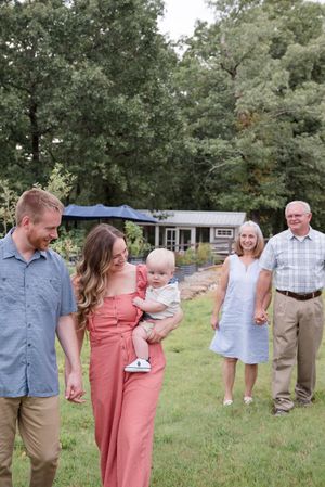 Family members hold hands while walking together on a grassy lawn surrounded by trees.