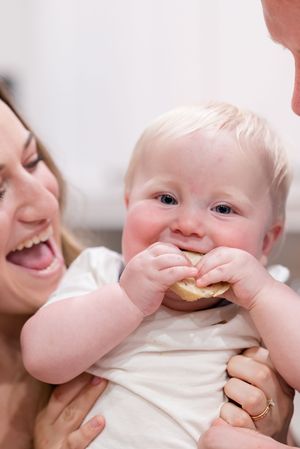Happy baby eating a snack while being held and smiling with rosy cheeks.