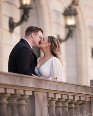 Romantic couple sharing an intimate moment on an elegant balcony with ornate stone columns and vintage wall lamps.