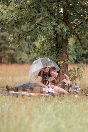 A clear umbrella provides shelter for a couple relaxing on a blanket under a leafy tree.