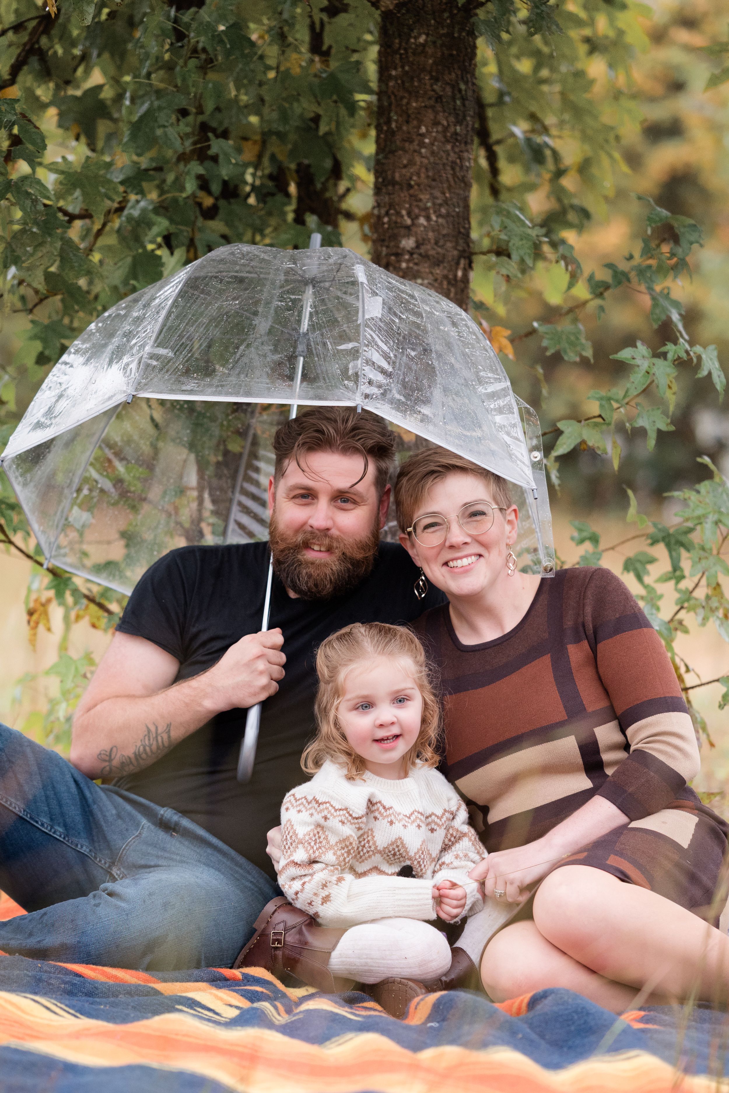Family relaxing together on an orange blanket under a tree with clear umbrellas.