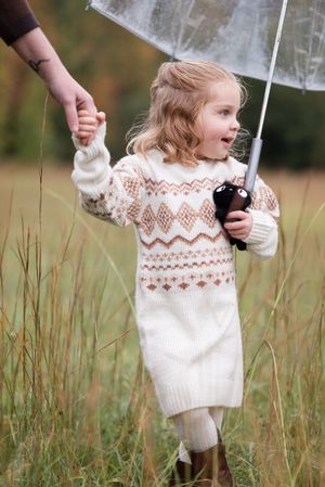 A child in a patterned white sweater dress holds a clear umbrella while walking through tall grass in autumn.