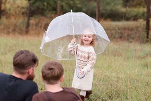 Family members share playful moments under a clear umbrella in a grassy field on an autumn day.