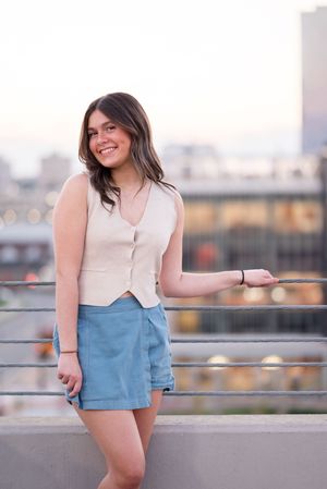 A person wearing a white button-up top and light blue shorts poses against a railing with a city skyline in the background.