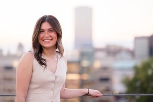 Person in a beige sleeveless top stands on urban rooftop balcony with blurred city skyline in background at dusk.