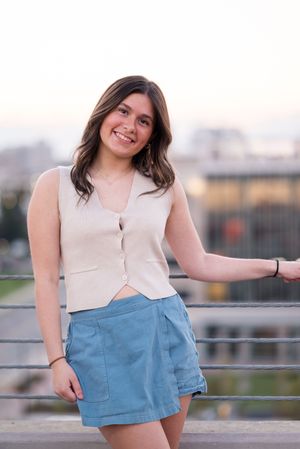 A model poses outdoors wearing a white sleeveless top and light blue shorts against an urban backdrop.