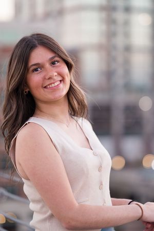 A person in a sleeveless cream top poses outdoors with city buildings in the background.