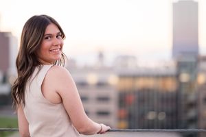 Woman in sleeveless top smiles while posing against blurred city skyline at sunset.